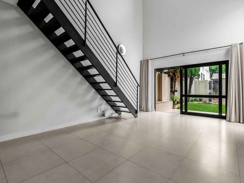 Spacious open-plan living area featuring high ceilings, modern tiled flooring, a striking metal staircase, and direct access to a private garden through sliding doors.