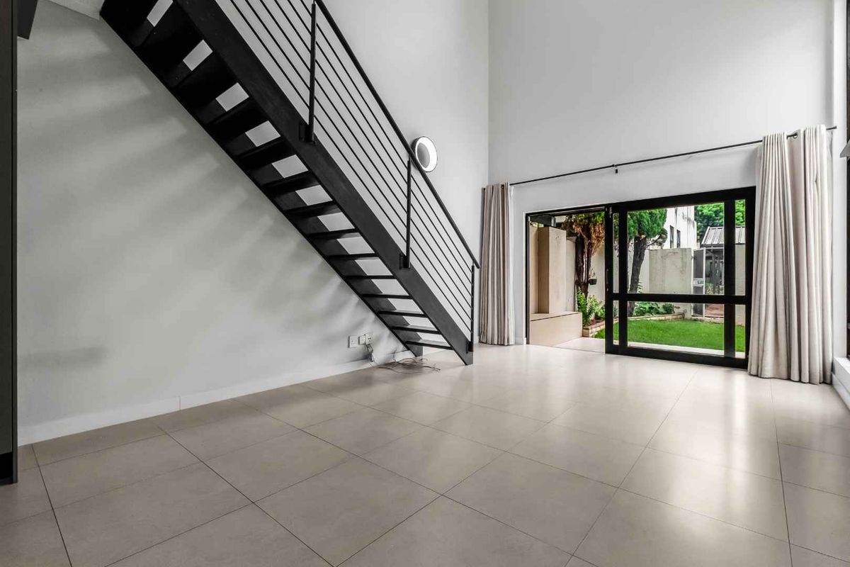 Spacious open-plan living area featuring high ceilings, modern tiled flooring, a striking metal staircase, and direct access to a private garden through sliding doors.