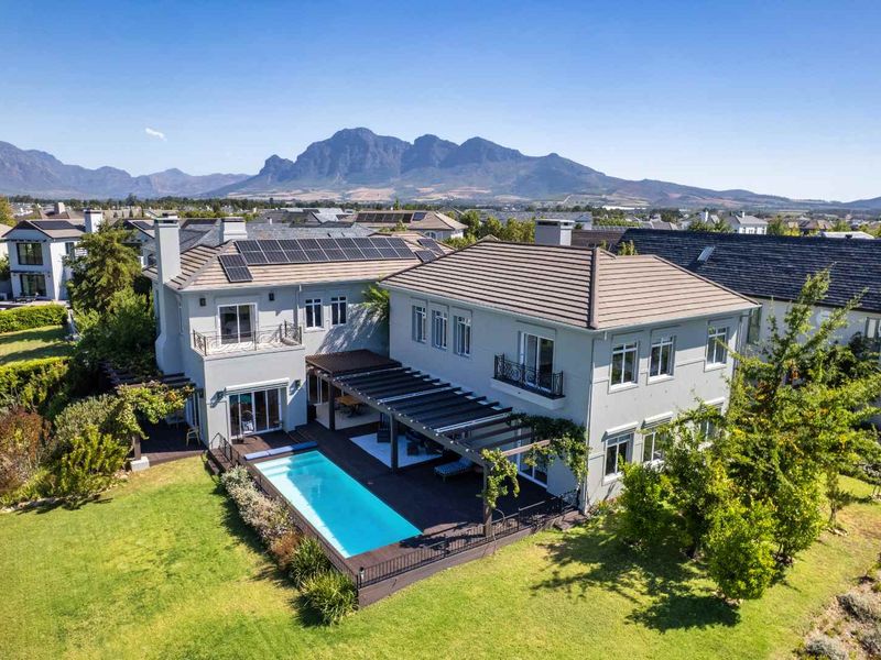 Aerial view of a two-story residence featuring a swimming pool, wooden deck, pergola, and solar panels, set against a scenic mountain backdrop.