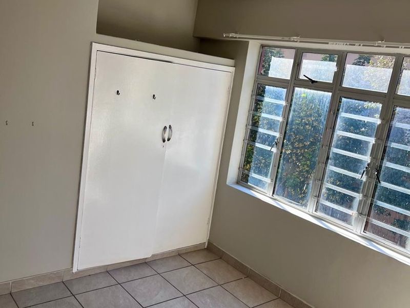 An empty bedroom featuring tiled flooring, a built-in cupboard, a ceiling fan, and large windows with security bars.