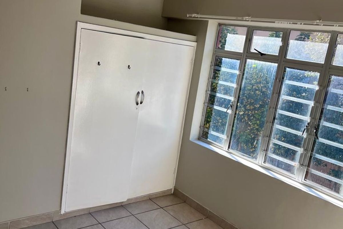 An empty bedroom featuring tiled flooring, a built-in cupboard, a ceiling fan, and large windows with security bars.