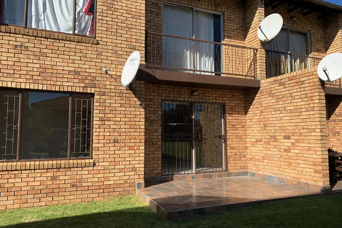 Rear view of a face-brick townhouse featuring a tiled patio, a small balcony with metal railing, and security gates on ground floor access points.