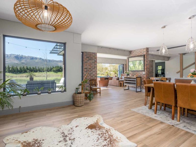 Spacious open-plan living and dining area featuring exposed brick accents, timber flooring, and large windows with scenic views and seamless indoor-outdoor flow.