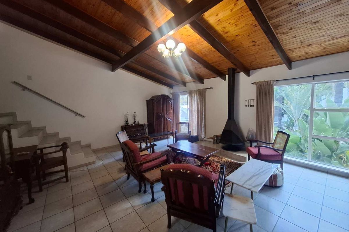 Spacious living room featuring a vaulted timber-beamed ceiling, tiled flooring, a freestanding fireplace, and large windows overlooking the garden.