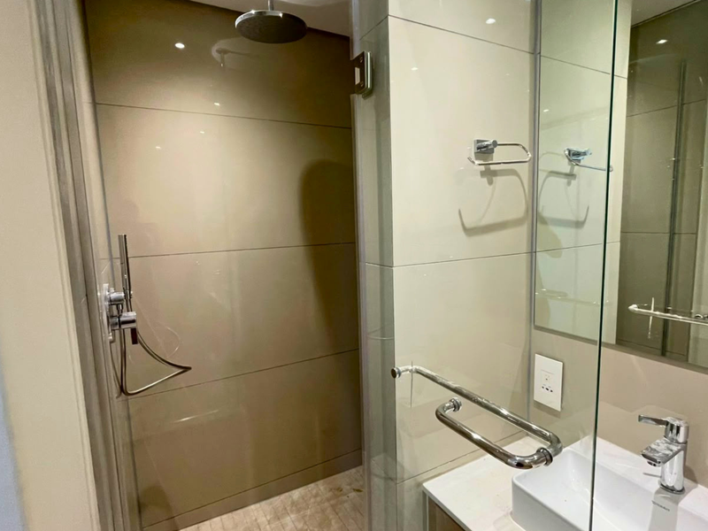 Modern bathroom featuring a glass-enclosed rain shower, wall-mounted mirror, vanity basin with chrome fixtures, and neutral tiled finishes.