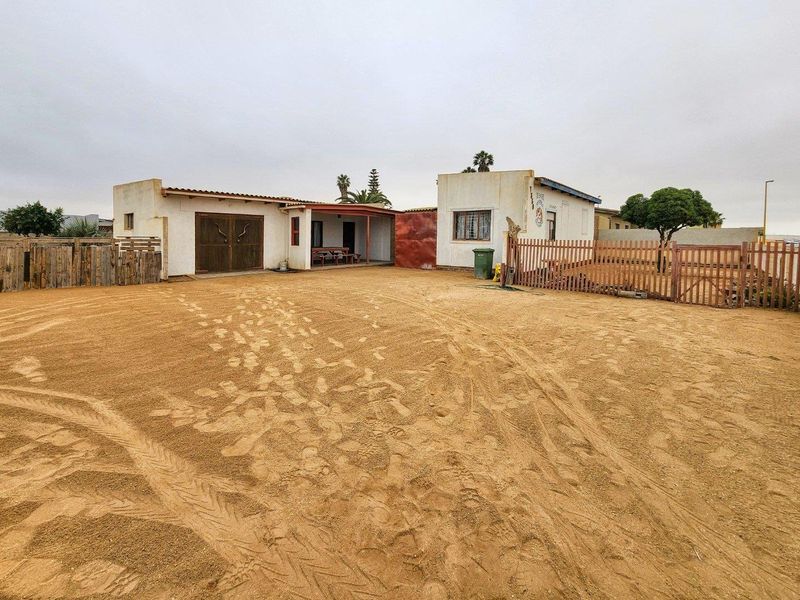 Front view of a single-story house featuring a covered patio, wooden garage doors, and a large sandy driveway area enclosed by wooden fencing.
