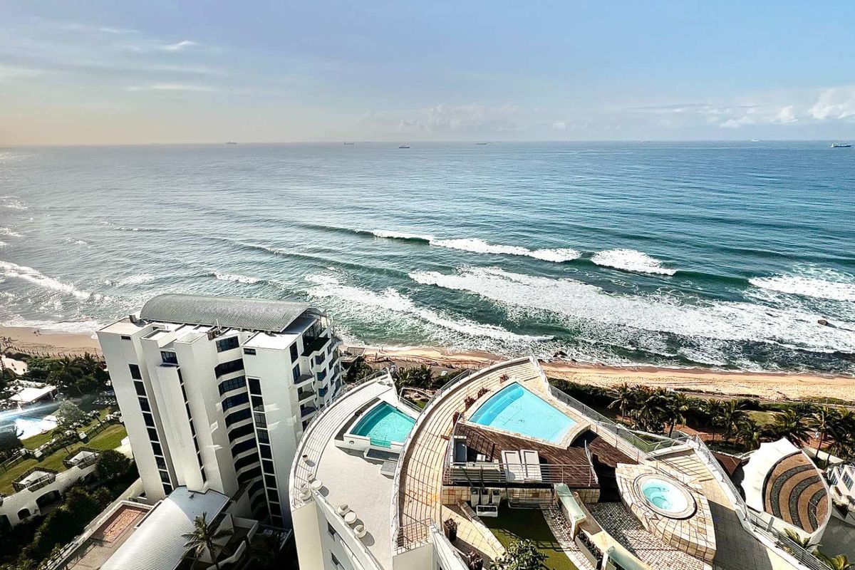 An elevated aerial view showcasing the apartment complex, featuring rooftop swimming pools, a jacuzzi, and direct oceanfront access.