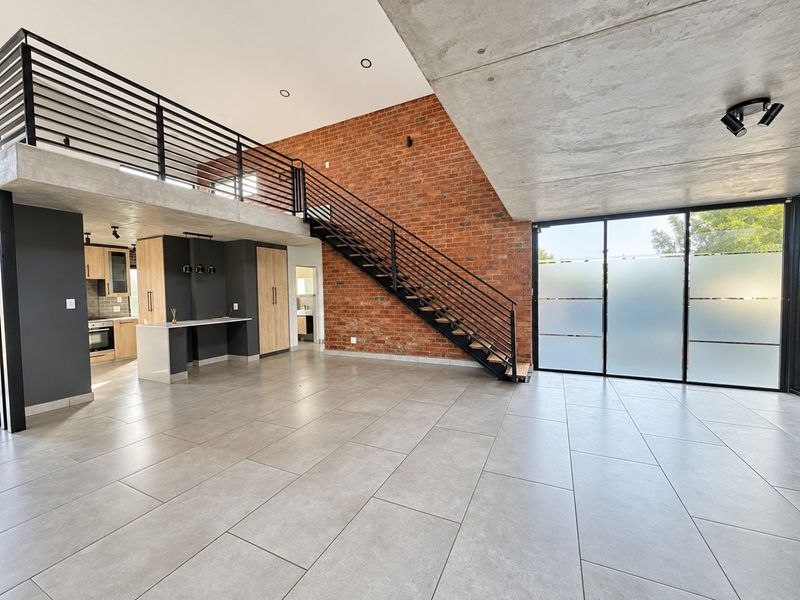 Spacious open-plan living area featuring industrial design elements, exposed brickwork, a concrete ceiling, and a modern staircase leading to a mezzanine level.
