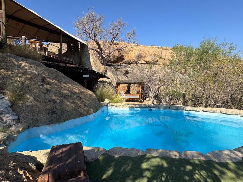 Outdoor swimming pool area integrated into natural rock formations, featuring a canvas shade structure and stone coping in a scenic lodge setting.