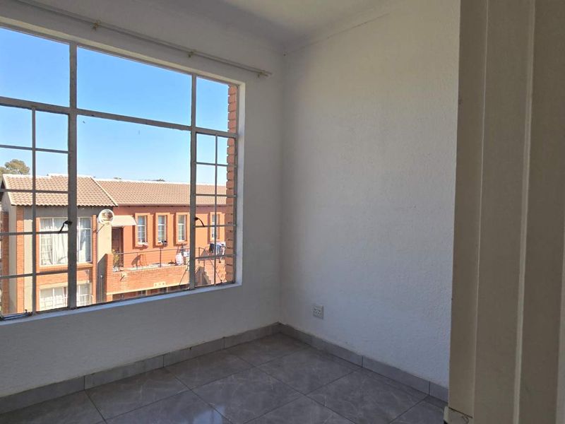 A bright, empty bedroom featuring tiled flooring and a large window providing natural light and a view of the surrounding complex.