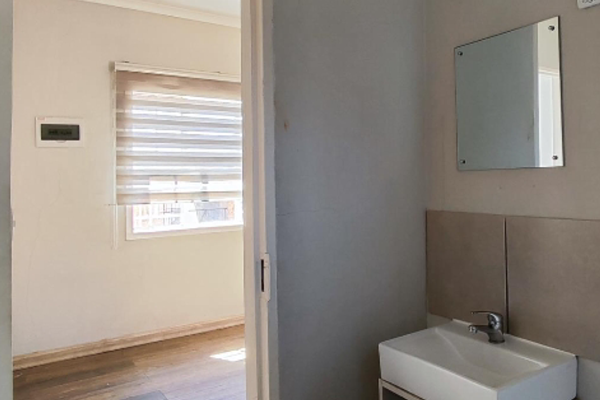 A compact bathroom vanity area featuring a modern basin and wall-mounted mirror, with a view into an adjacent bedroom with wood-look flooring.
