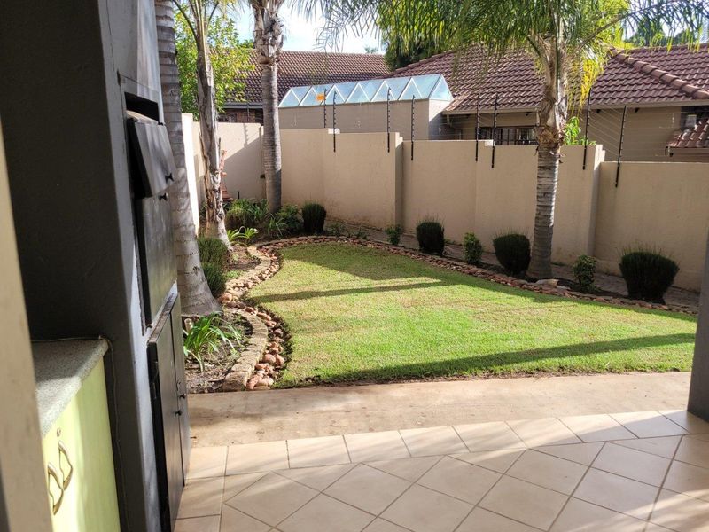 A private rear garden featuring a tiled patio, built-in braai, manicured lawn, and secure perimeter wall with electric fencing.