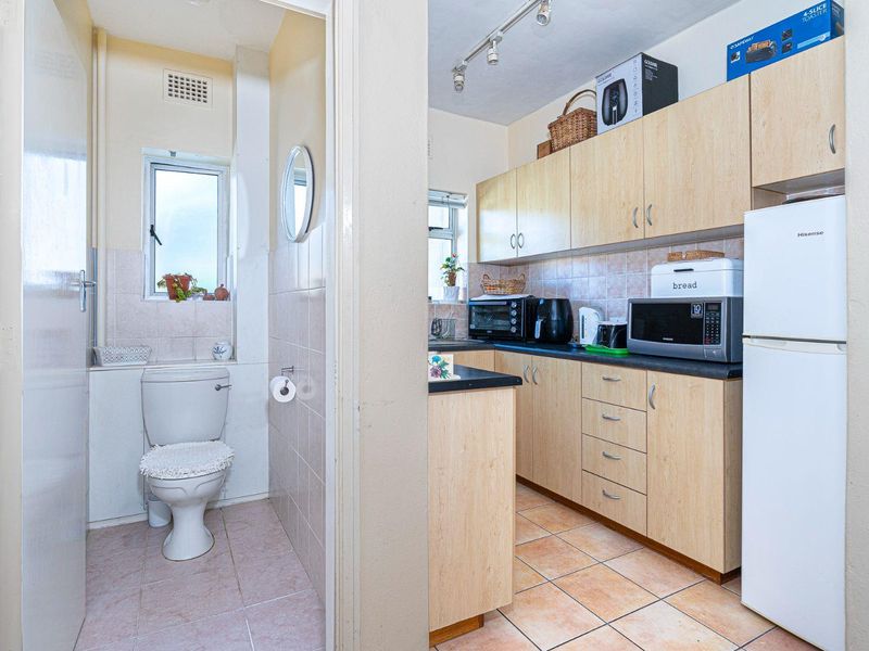 A functional kitchen featuring light wood-finish cabinetry and tiled flooring, adjacent to a compact bathroom with a standard toilet and tiled finishes.