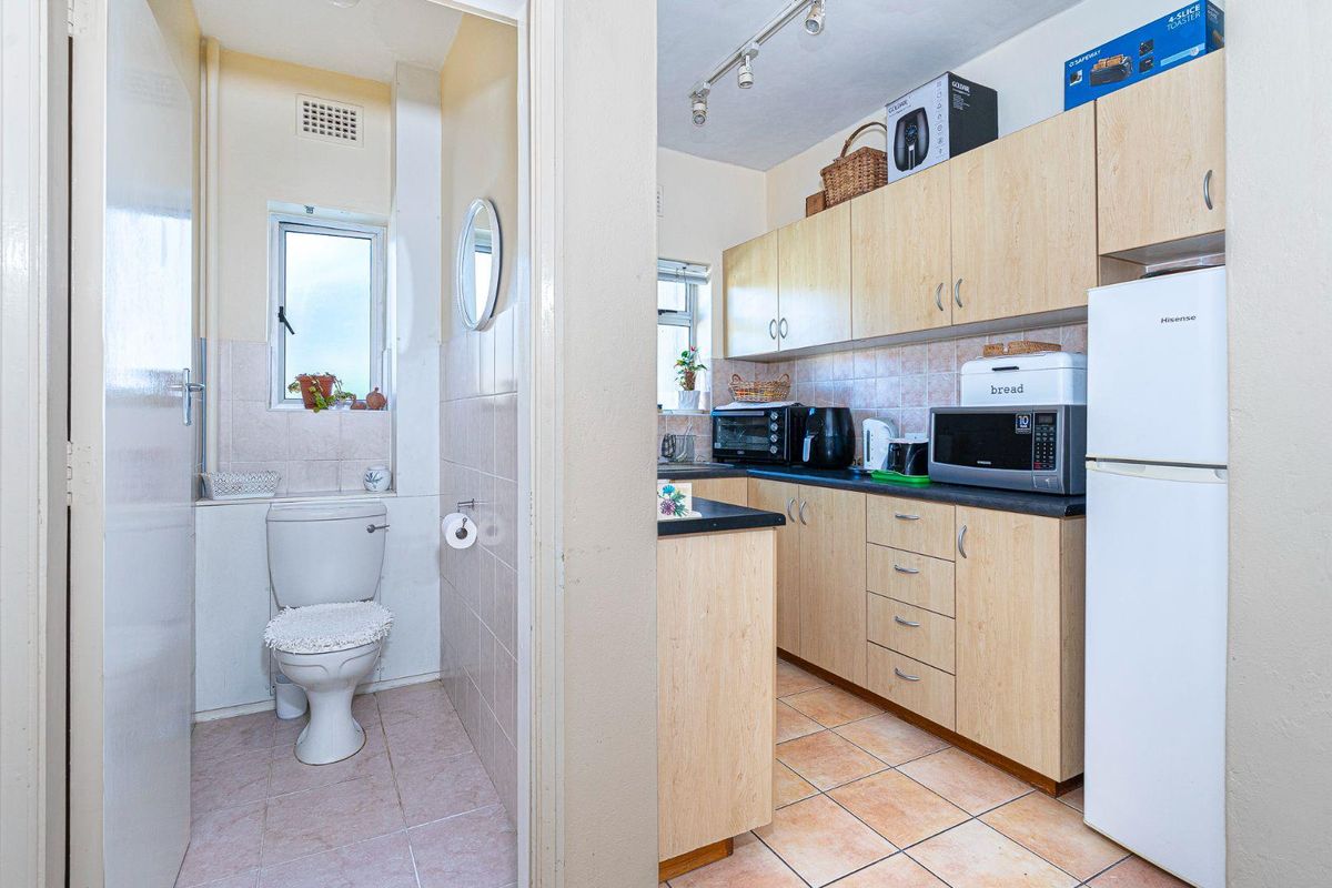 A functional kitchen featuring light wood-finish cabinetry and tiled flooring, adjacent to a compact bathroom with a standard toilet and tiled finishes.