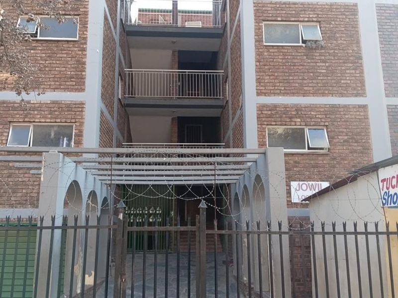 Front elevation of a multi-story apartment building featuring face brick construction, metal security fencing with razor wire, and private balconies.