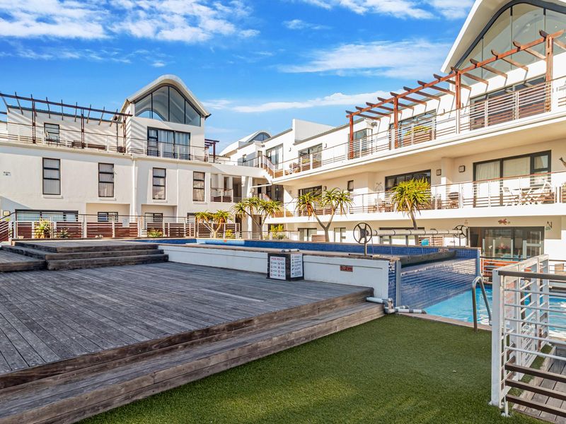 Communal pool area featuring a swimming pool, timber decking, and artificial lawn set within a modern apartment complex.