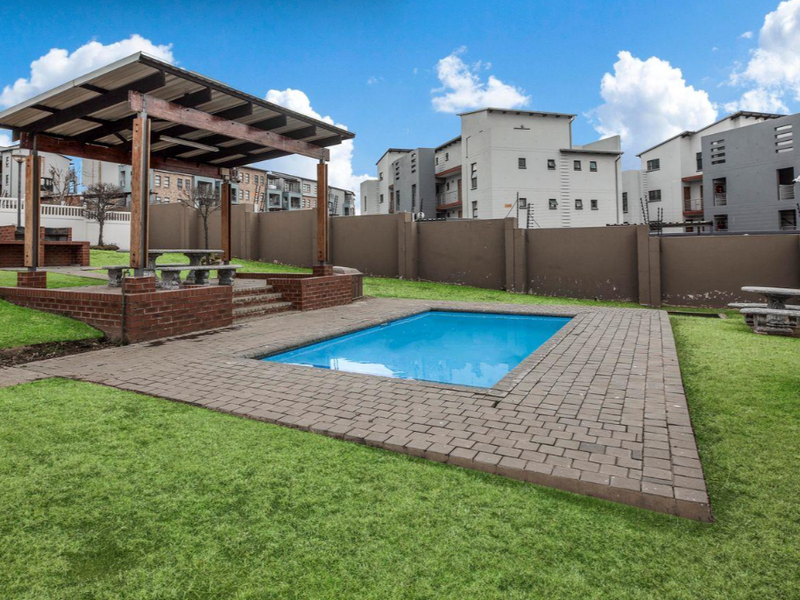 Communal outdoor pool area featuring a swimming pool, brick-paved surround, timber pergola, and a built-in brick braai facility within a secure complex.