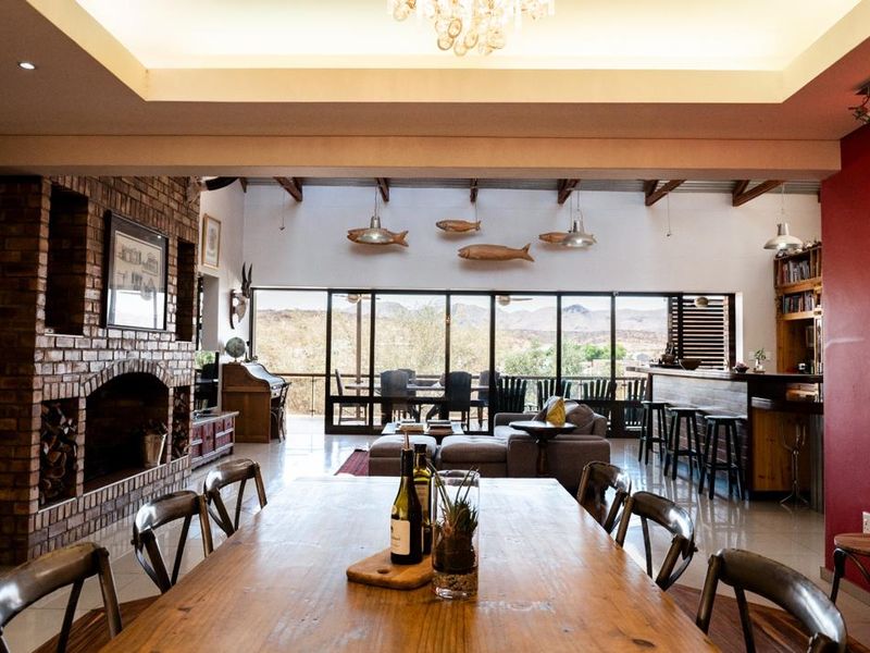 Spacious open-plan living and dining area featuring an exposed brick fireplace, high ceilings with timber beams, and large sliding doors opening to a scenic view.