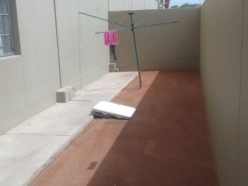A narrow rear exterior area featuring concrete paving, a perimeter wall with security fencing, and an unpaved ground section.