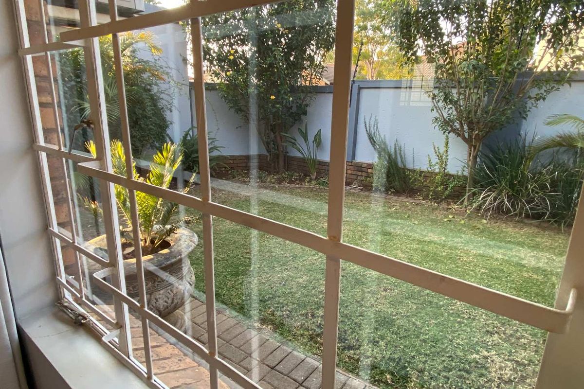 A view of the private rear garden featuring a paved walkway, lawn, and perimeter brick boundary wall, viewed through a window with security bars.