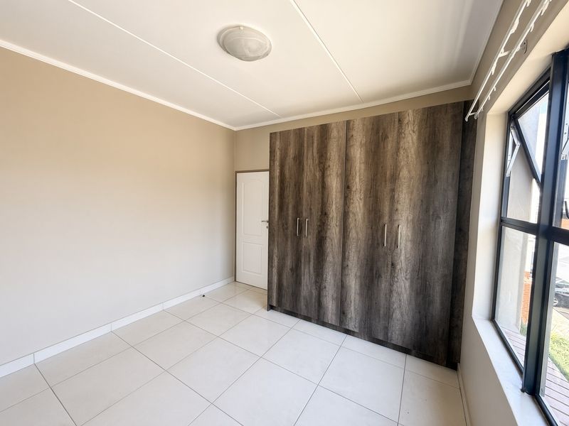A bright, empty bedroom featuring modern wood-look built-in cupboards, neutral tiled flooring, and large windows providing natural light.