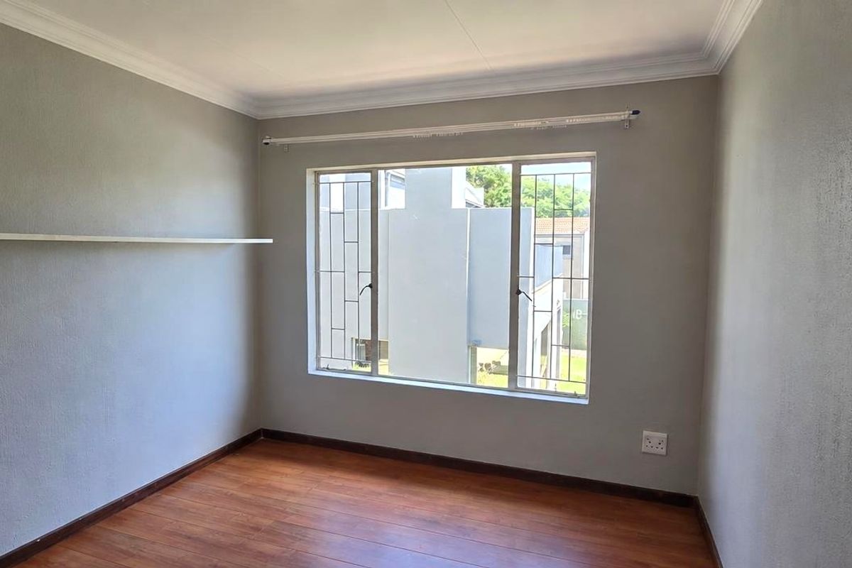 A bright, empty bedroom featuring laminate flooring, neutral grey walls, a wall-mounted shelf, and a window fitted with security bars.