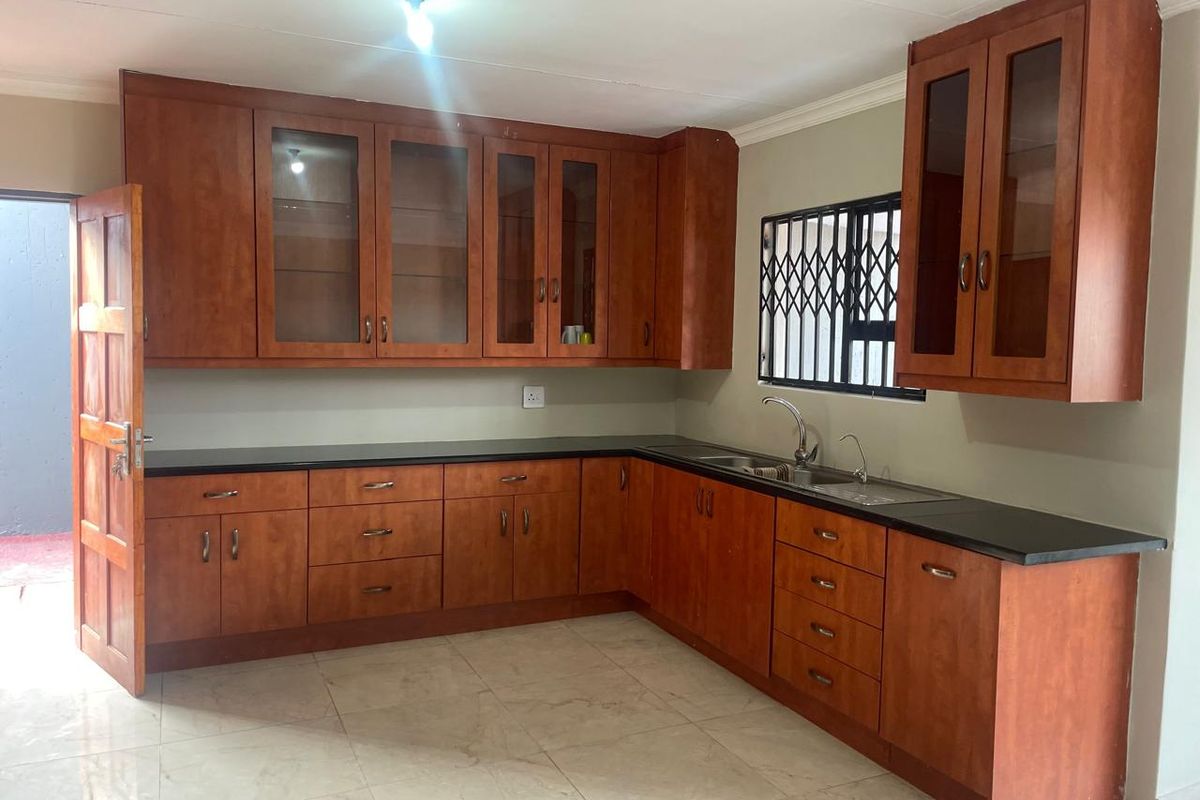 A functional kitchen featuring wooden cabinetry, dark countertops, a double sink, and tiled flooring with a security gate on the window.