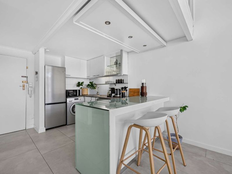 Modern compact kitchen featuring white cabinetry, a glass-topped breakfast bar, tiled flooring, and integrated appliances including a washing machine and refrigerator.