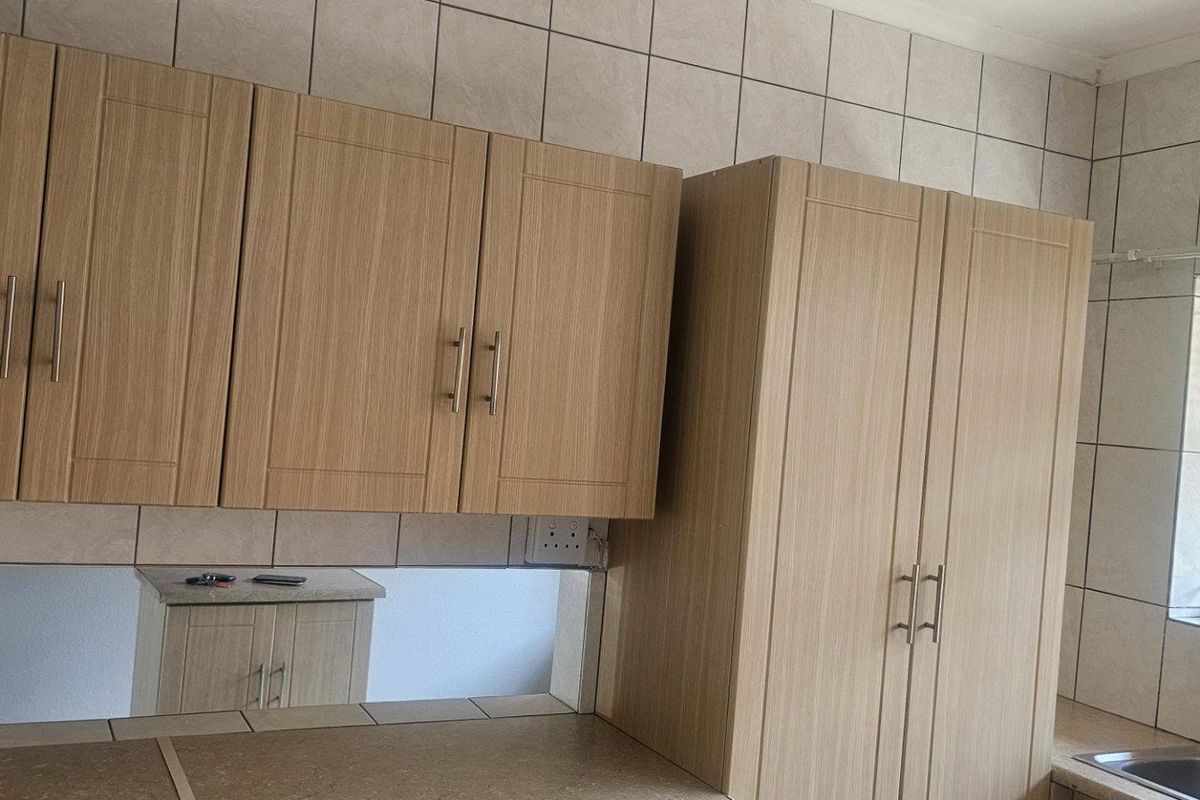A functional kitchen area featuring light wood-finish cabinetry, tiled splashbacks, a stainless steel sink, and a window with security bars overlooking the garden.