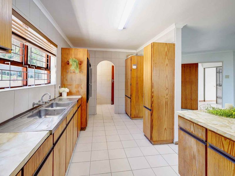 A functional kitchen featuring wooden cabinetry, double stainless steel sink, tiled flooring, and a neutral tiled splashback, offering a clean and practical layout.