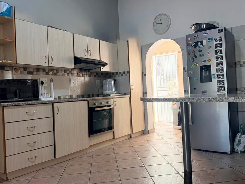 A functional kitchen featuring light wood-finish cabinetry, granite countertops, built-in oven, extractor fan, and tiled flooring, leading through an archway to an exterior area.