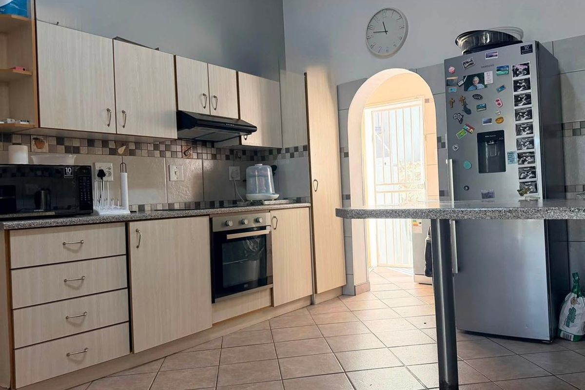 A functional kitchen featuring light wood-finish cabinetry, granite countertops, built-in oven, extractor fan, and tiled flooring, leading through an archway to an exterior area.