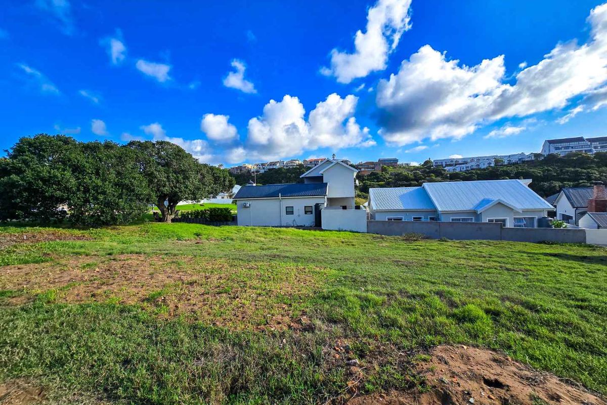 A vacant residential plot of land featuring established grass, mature trees, and a clear view of neighboring properties under a bright blue sky.
