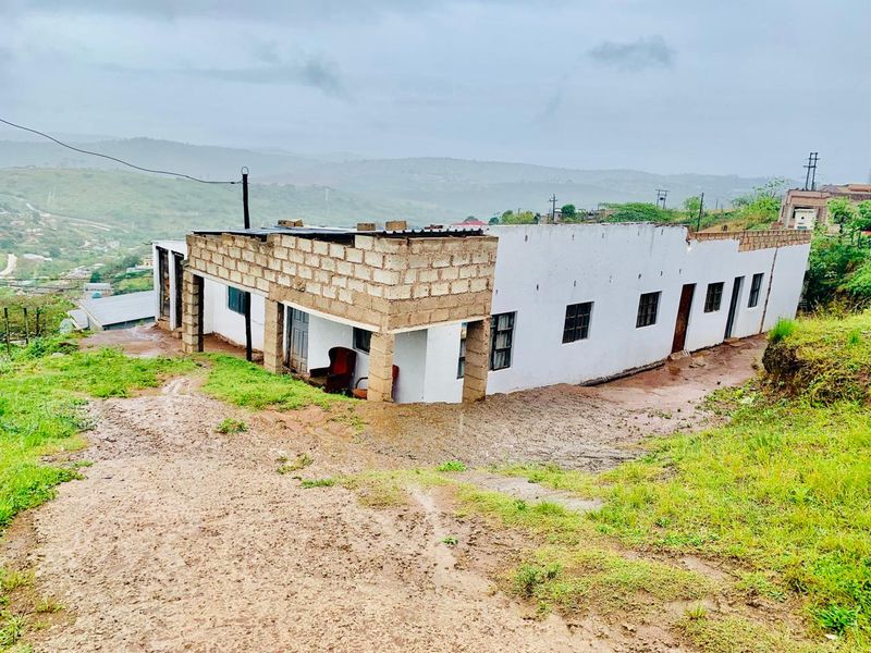 Exterior view of a single-story house featuring unfinished concrete block walls and a corrugated metal roof, situated on an unpaved, sloped site.
