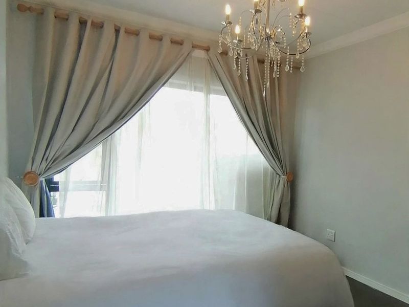 A well-lit bedroom featuring a decorative chandelier, neutral wall finishes, and large window with curtain rod.