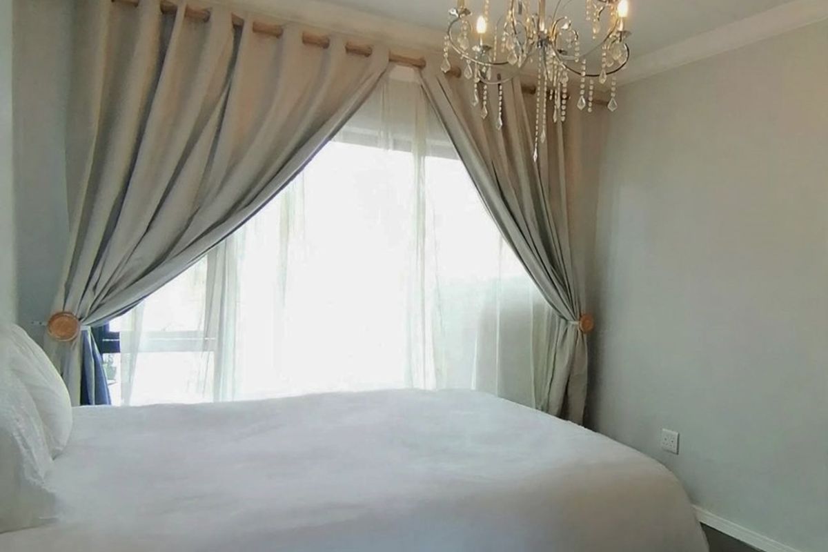 A well-lit bedroom featuring a decorative chandelier, neutral wall finishes, and large window with curtain rod.