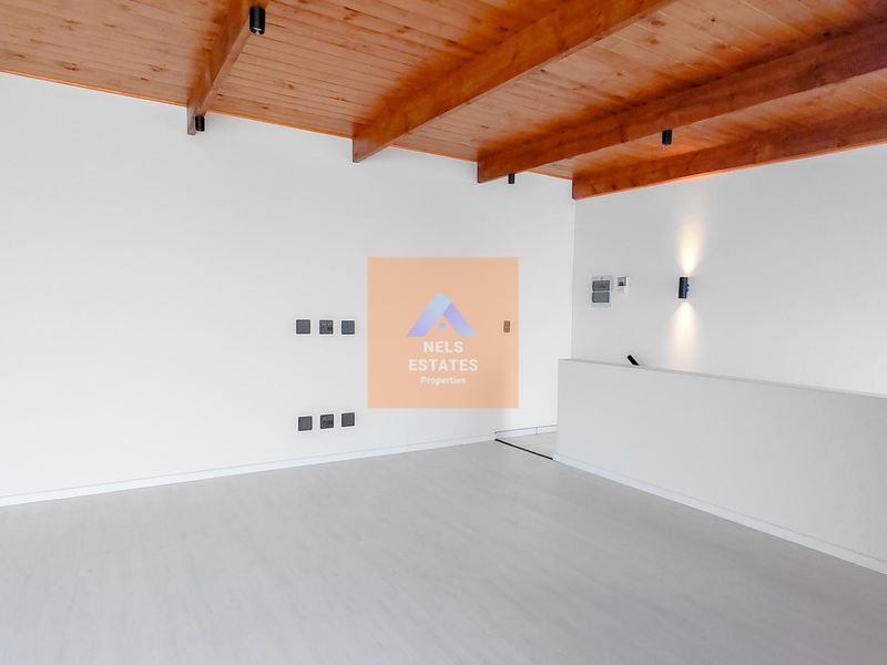 A bright, empty living area featuring exposed timber ceiling beams, neutral flooring, and modern wall-mounted lighting, offering a clean canvas for interior design.