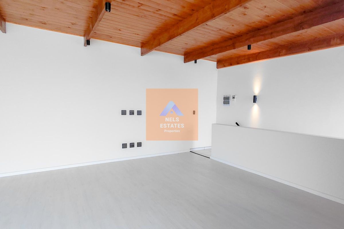 A bright, empty living area featuring exposed timber ceiling beams, neutral flooring, and modern wall-mounted lighting, offering a clean canvas for interior design.