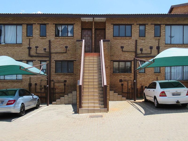 Front elevation of a face brick apartment complex featuring a central staircase, paved parking area, and dedicated carport shade structures.
