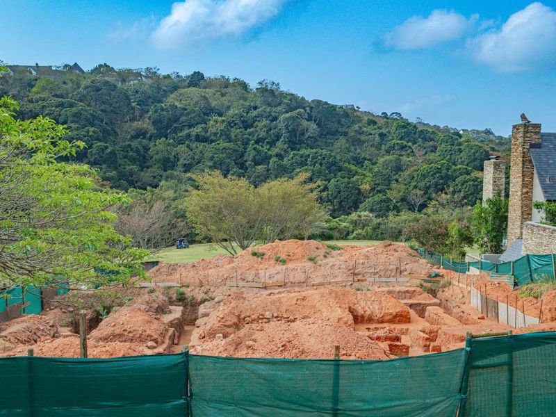 A view of a residential vacant land plot showing an active construction site with excavated foundations and perimeter fencing against a natural landscape backdrop.