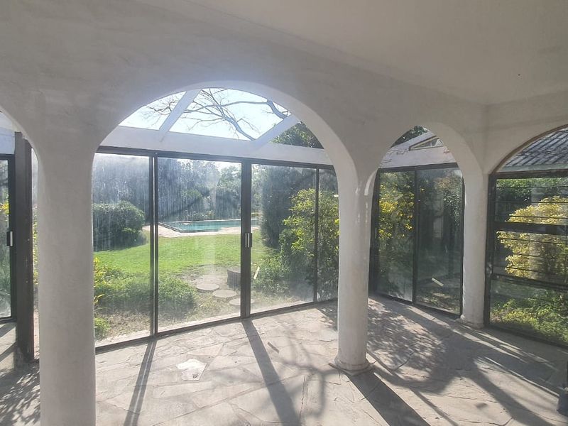 A covered patio area featuring stone flooring, arched columns, and glass sliding doors overlooking the garden and pool area.