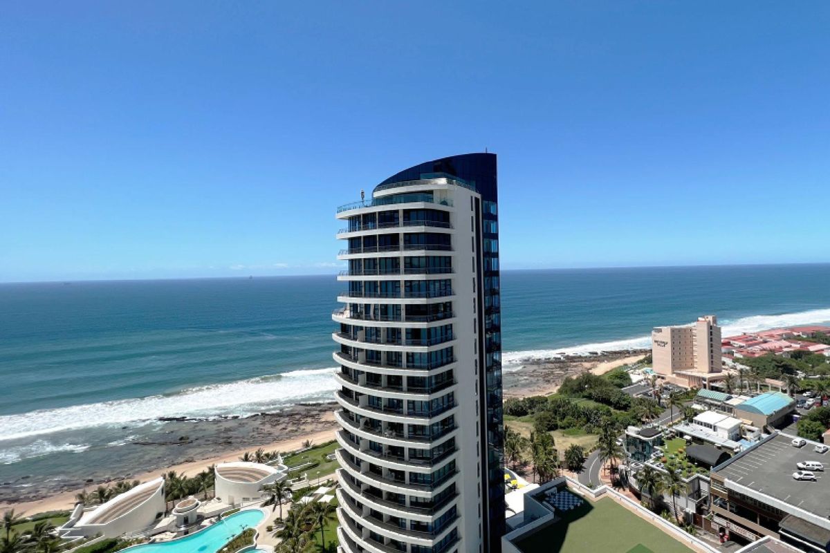 Aerial view of a modern high-rise apartment building situated along the coastline, featuring balconies and access to a swimming pool and landscaped grounds.
