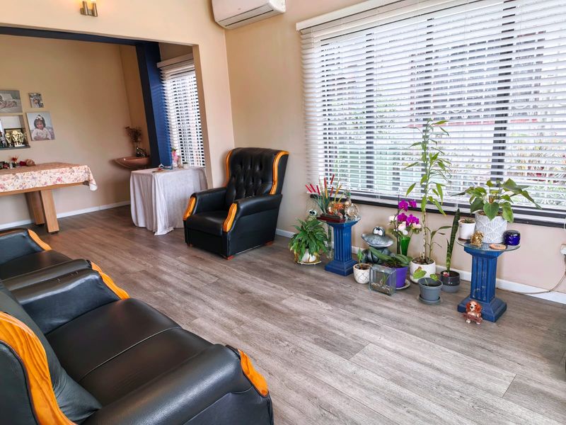 A living room featuring wood-look laminate flooring, venetian blinds, and a wall-mounted air conditioning unit, with an adjacent alcove containing a small basin.