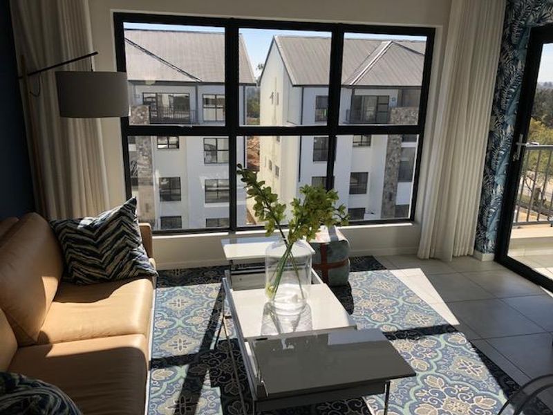A bright living room featuring large aluminium-framed windows, tiled flooring, and direct access to an outdoor balcony area.
