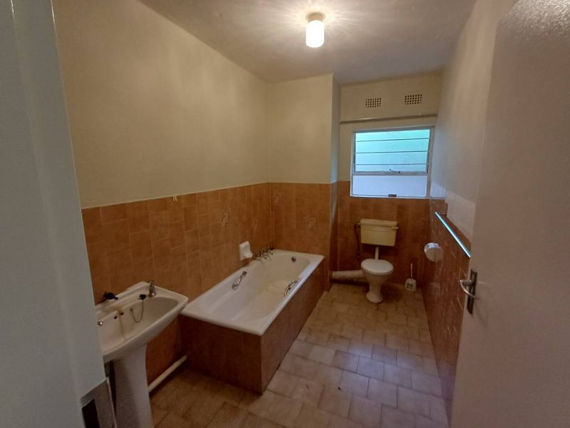 A dated bathroom featuring a pedestal basin, built-in bathtub, and toilet, finished with original ceramic wall tiles and louvered window.