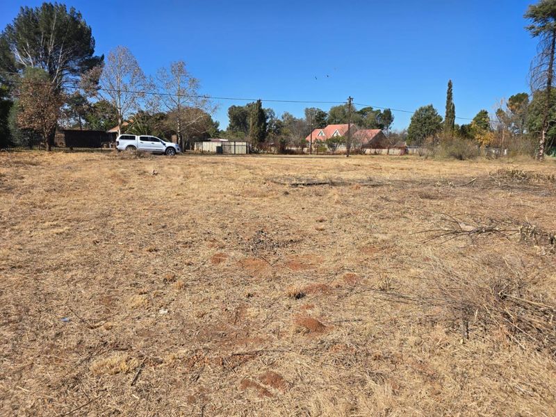 A vacant residential plot of land featuring dry grass, mature trees, and existing perimeter fencing, suitable for future development.