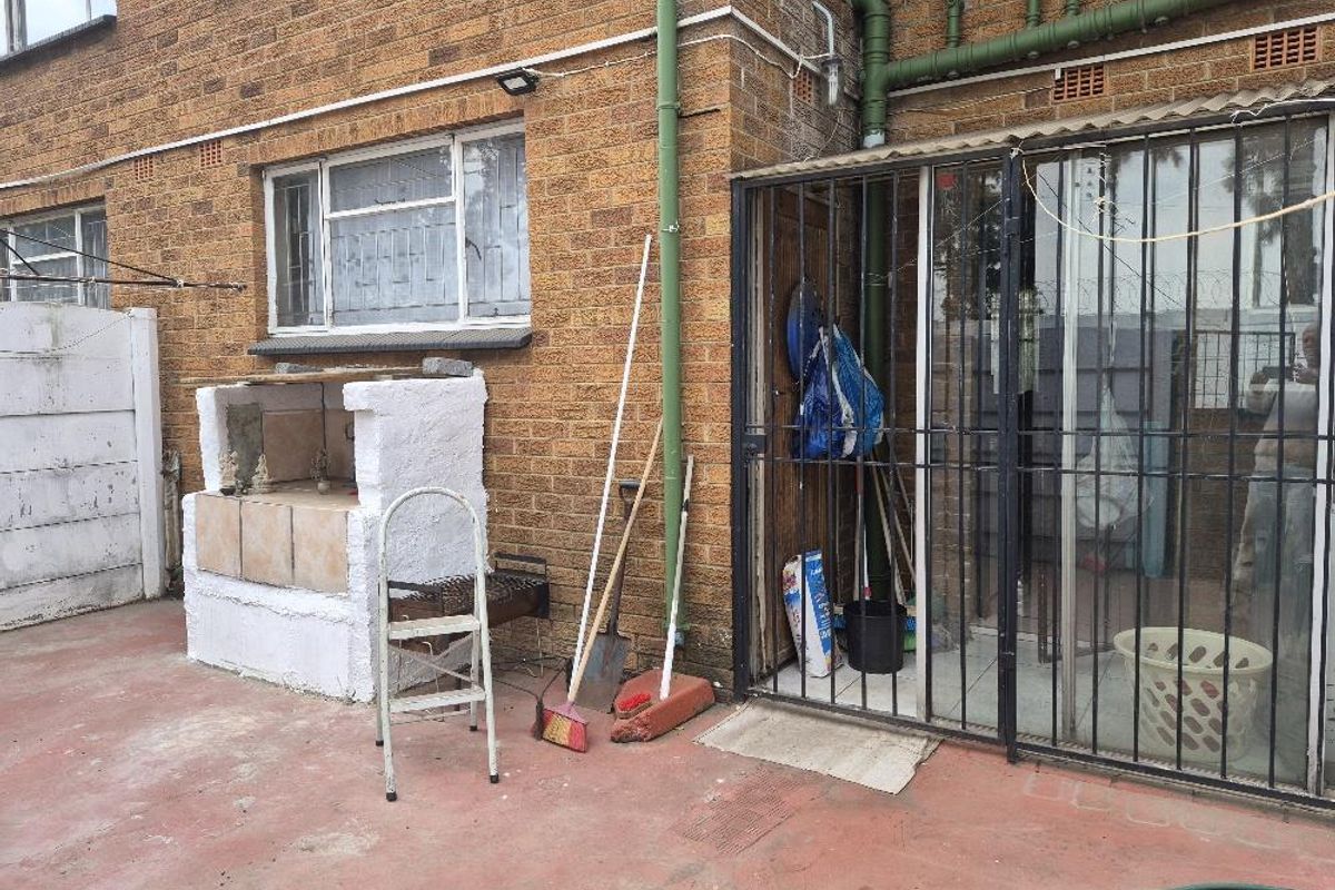 Rear exterior area featuring a built-in brick braai, security gate access, and paved flooring, showing signs of age and wear.