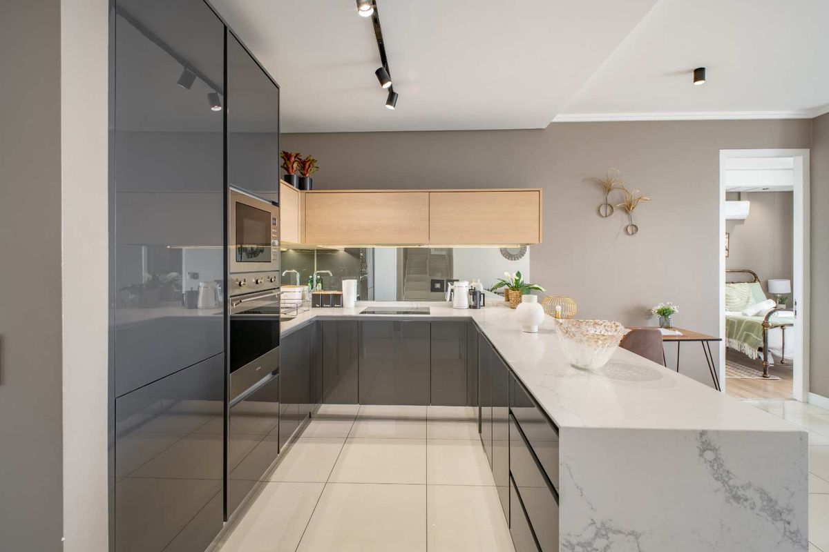 Modern kitchen featuring high-gloss grey cabinetry, light wood accents, stone countertops, integrated oven, and track lighting, seamlessly flowing into the adjacent living area.