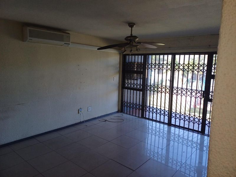 An empty living room featuring tiled flooring, a ceiling fan, a wall-mounted air conditioner, and a security gate leading to an outdoor area.