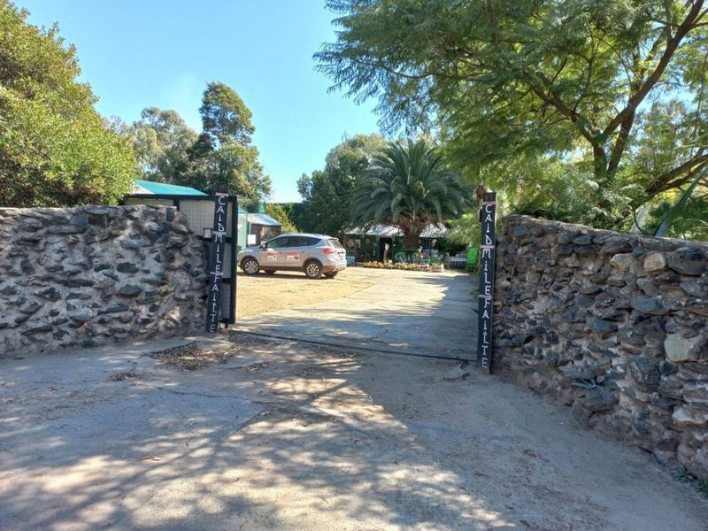 Entrance view of the small holding property featuring a stone boundary wall, concrete driveway, and mature garden landscaping.